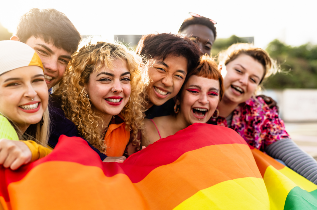 A group photo of smiling young people holding a large rainbow flag.